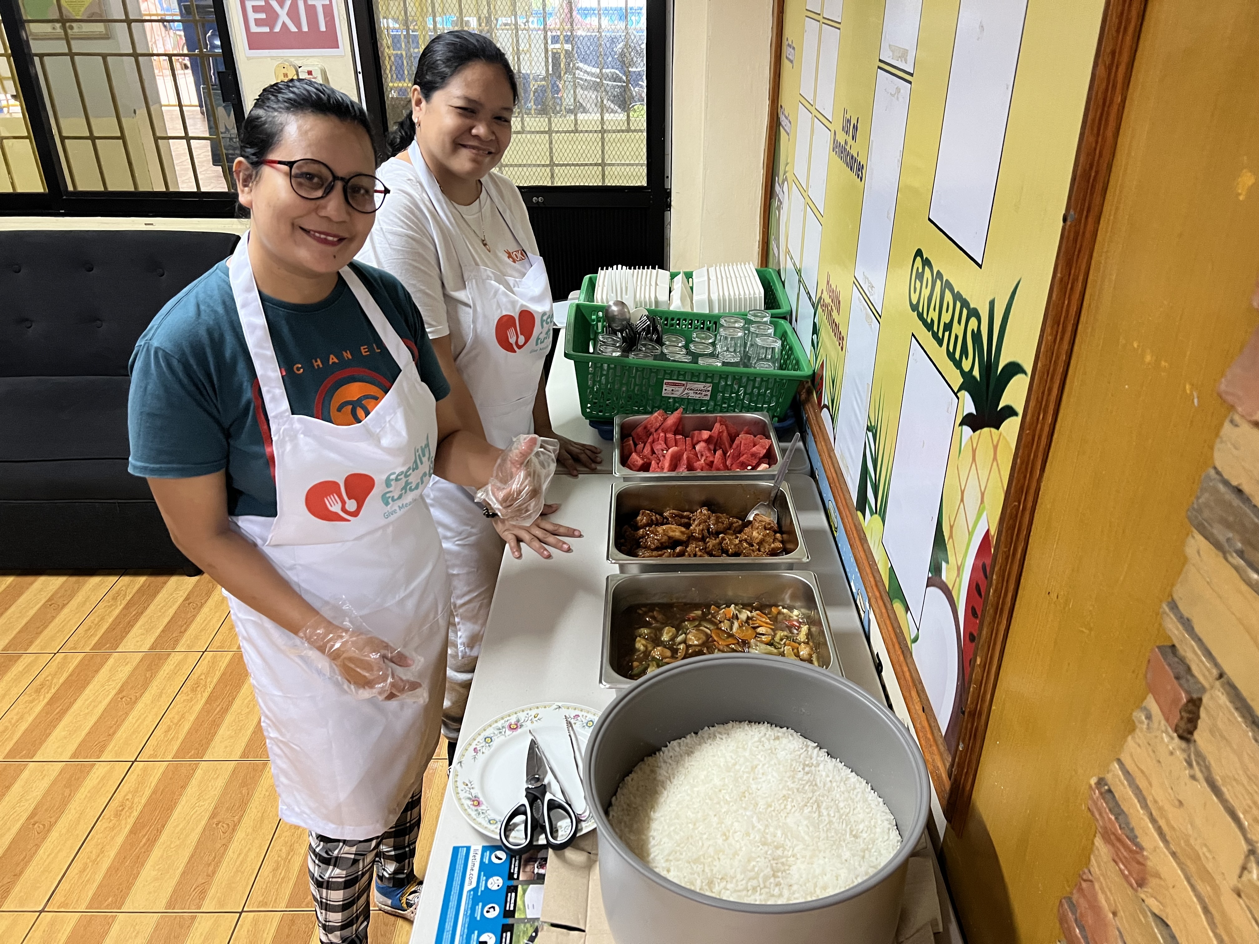 Two smiling volunteers at the serving station with trays of hot food.