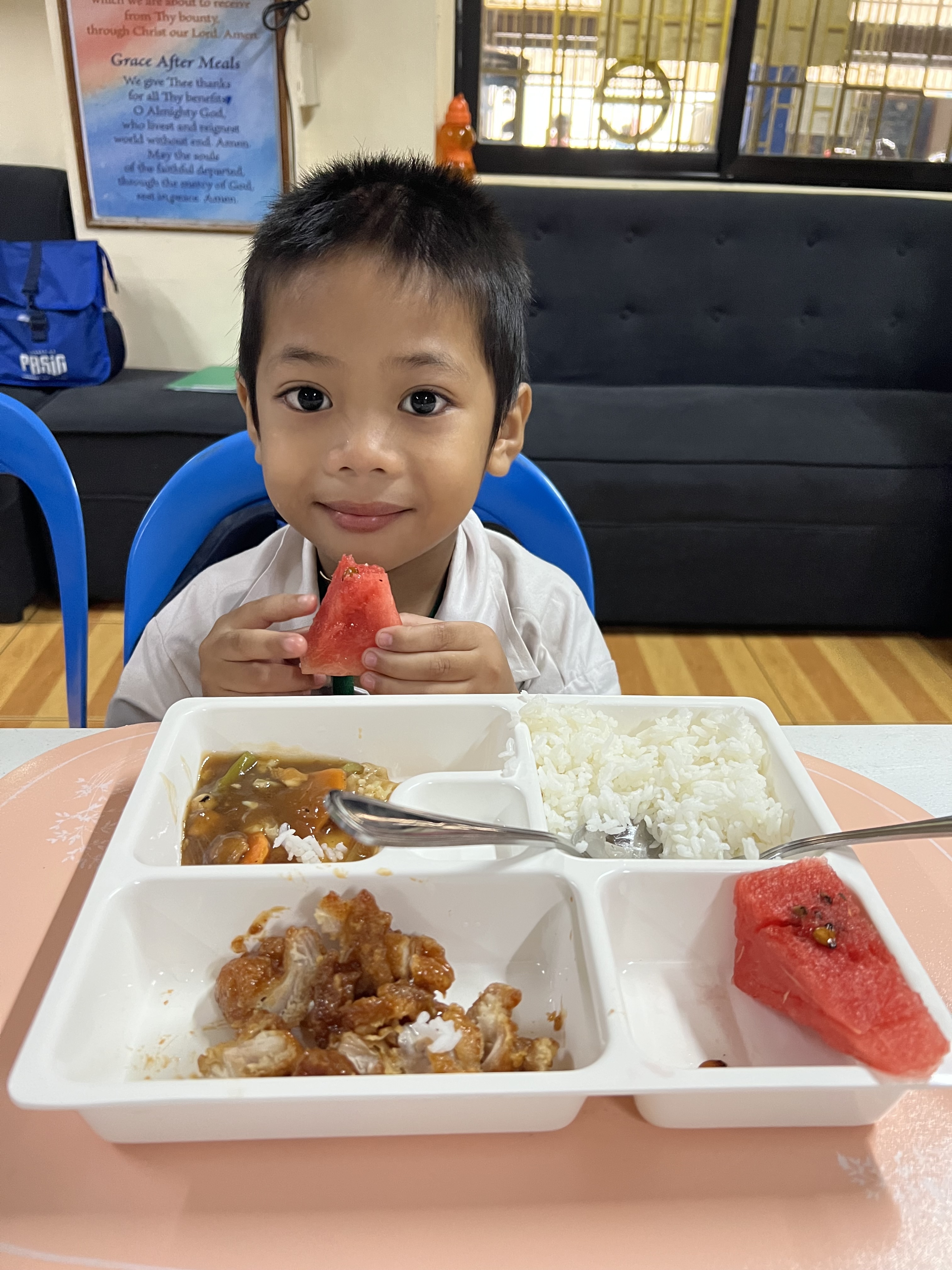 A young boy smiling at the camera while holding a piece of watermelon from his meal.